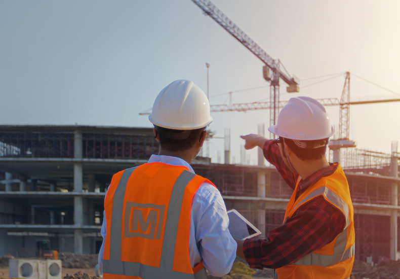Bauingenieure mit Schutzhelmen und Tablets koordinieren Tragwerksplanung auf einer Baustelle mit sichtbaren Kränen und Rohbau.